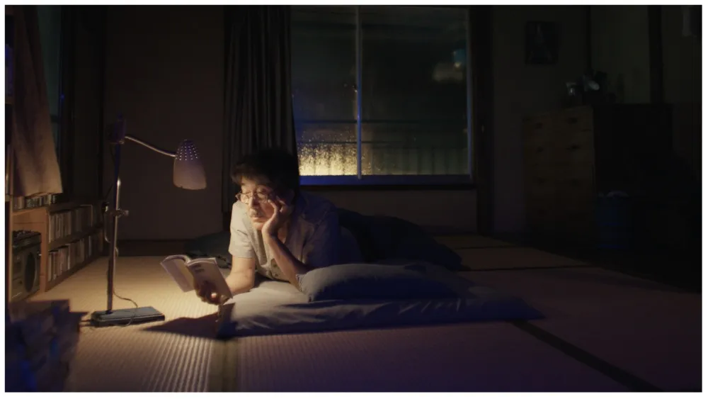 Perfect Days filminden bir karakter, sıradan gözüken bir evde, gece karanlığında kitap okuyor