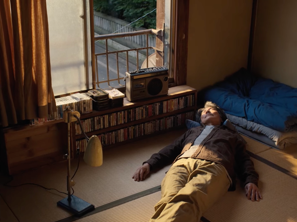 Perfect Days filminden bir karakterin üç sıra boyunca dizilmiş müzik kasetlerinin yanında yere uzanarak müzik dinlediği bir görsel